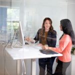 Two colleagues in a one-on-one conversation at a white standing desk in a glass-walled office, one gesturing toward a monitor while the other listens.