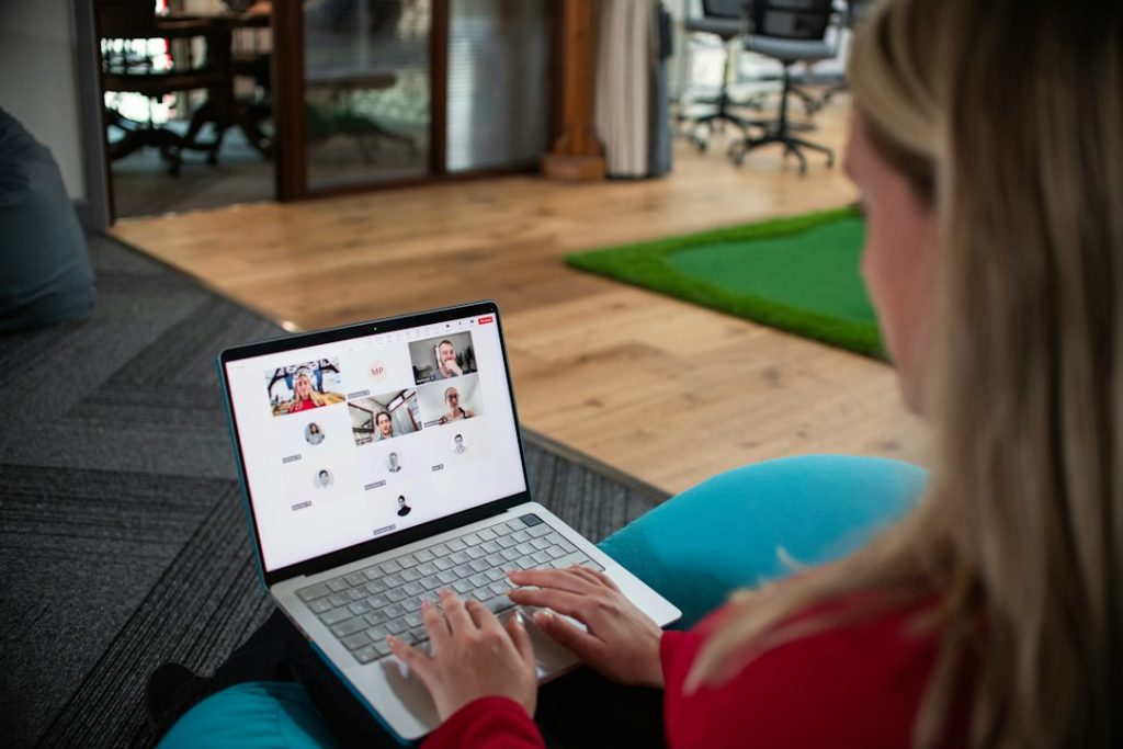 Over-the-shoulder view of a woman in a red top on a laptop, joined to a video call with a grid of participants, several tiles showing only initials or placeholders