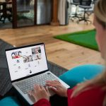 Over-the-shoulder view of a woman in a red top on a laptop, joined to a video call with a grid of participants, several tiles showing only initials or placeholders