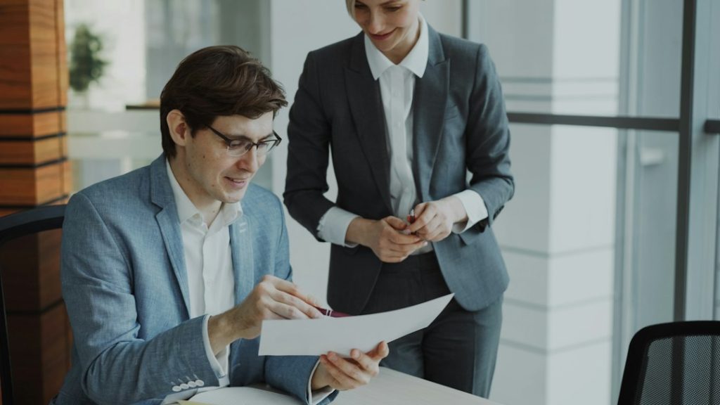 A seated man in a light blue blazer and glasses reads a printed page while a woman in a grey suit stands beside him, both smiling, in a bright office with glass walls