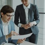 A seated man in a light blue blazer and glasses reads a printed page while a woman in a grey suit stands beside him, both smiling, in a bright office with glass walls