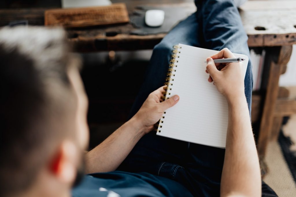 A person sitting with a blank spiral-bound notebook resting on one knee, pen in hand and poised to write, with a wooden table in the background.