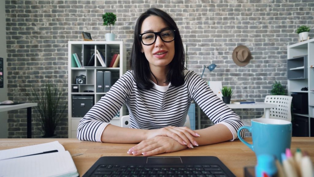 A woman in a black and white striped top sits at a wooden desk behind an open laptop, hands folded and looking directly into the camera as if speaking on a video call, in a home office with an exposed brick wall, white shelving and a notebook and mug on the desk.