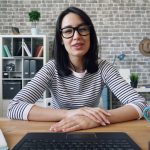 A woman in a black and white striped top sits at a wooden desk behind an open laptop, hands folded and looking directly into the camera as if speaking on a video call, in a home office with an exposed brick wall, white shelving and a notebook and mug on the desk.