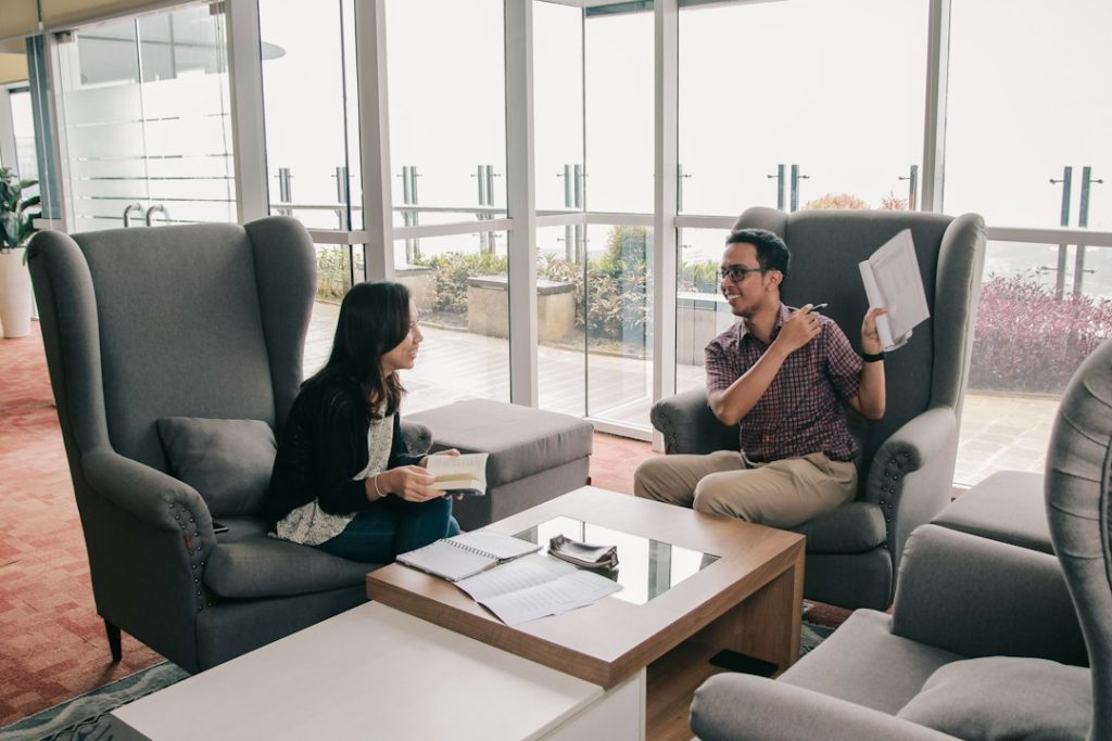 Two people sitting in grey wingback armchairs by a bright window, one holding an open notebook and pen mid-sentence, the other listening with a book in hand, papers and a spiral notebook on the coffee table between them