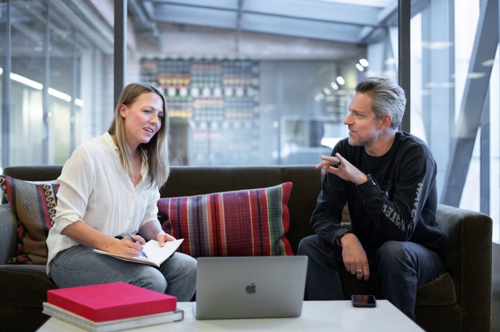 Two people sitting on a couch in a bright open-plan office having a one-on-one conversation, one taking handwritten notes in a notebook with an open laptop on the low table between them.