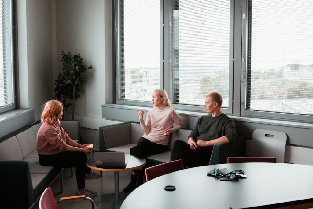 Three people in a bright office lounge talking, one gesturing as she speaks while the other two listen, with a laptop on a small table between them
