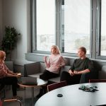 Three people in a bright office lounge talking, one gesturing as she speaks while the other two listen, with a laptop on a small table between them