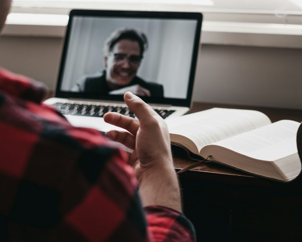 A coach in a red plaid shirt gestures during a video call with a smiling client on a laptop, an open book on the desk beside it.