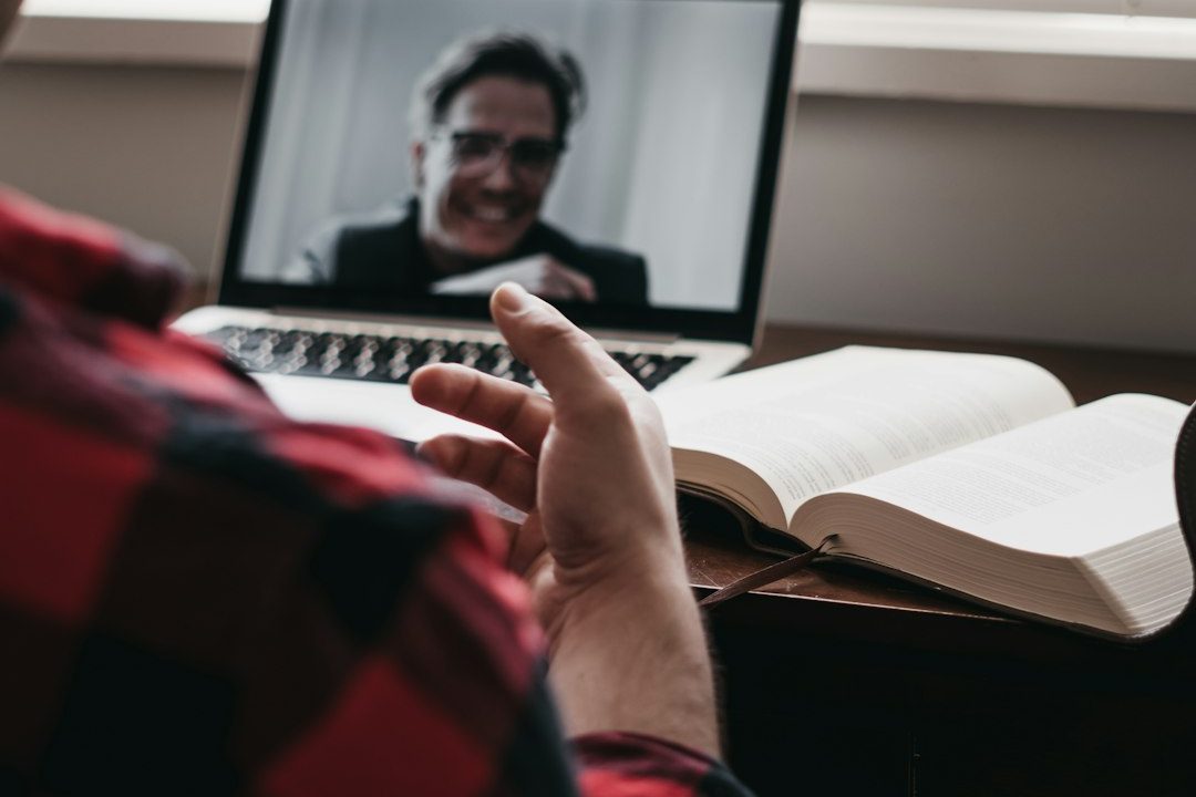 A coach in a red plaid shirt gestures during a video call with a smiling client on a laptop, an open book on the desk beside it.