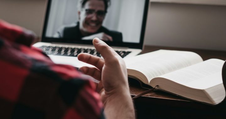 A coach in a red plaid shirt gestures during a video call with a smiling client on a laptop, an open book on the desk beside it.