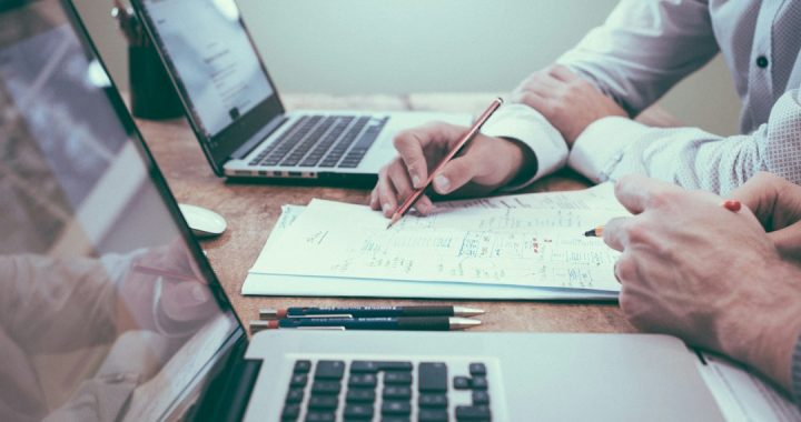 Two people at a wooden desk with laptops, one pointing a pencil at a page of handwritten planning notes while the other holds a pen