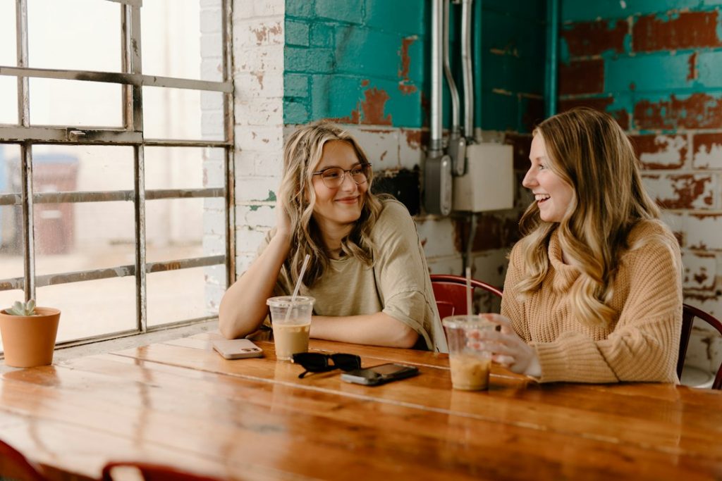 Two women talking and smiling across a wooden table in a bright cafe with iced coffees, a teal brick wall behind them