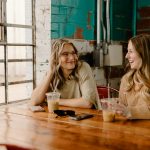 Two women talking and smiling across a wooden table in a bright cafe with iced coffees, a teal brick wall behind them