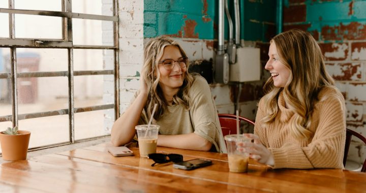 Two women talking and smiling across a wooden table in a bright cafe with iced coffees, a teal brick wall behind them