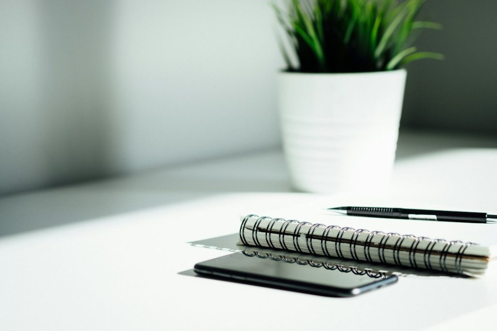 A spiral notebook, a black pencil and a phone lying face down on a white desk beside a potted plant in soft window light