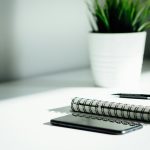 A spiral notebook, a black pencil and a phone lying face down on a white desk beside a potted plant in soft window light