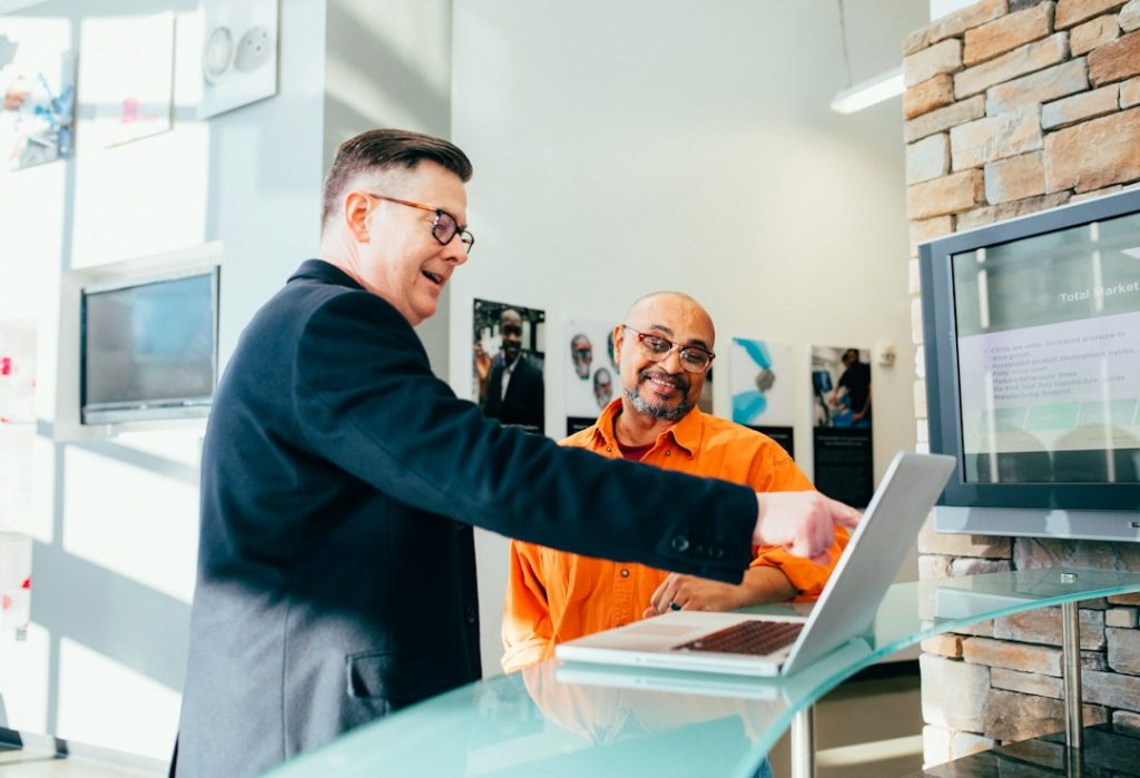 A man in a dark blazer and glasses smiles and points at an open laptop while a seated man in an orange shirt and glasses smiles back, in a bright office with a stone wall.