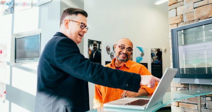 A man in a dark blazer and glasses smiles and points at an open laptop while a seated man in an orange shirt and glasses smiles back, in a bright office with a stone wall.