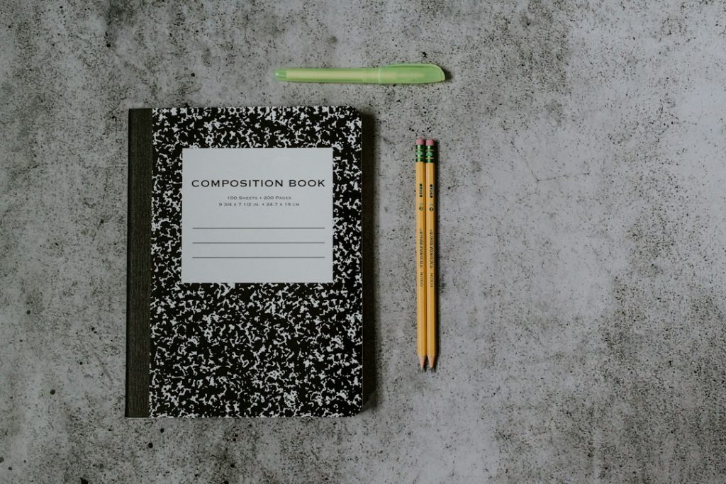 A black and white composition notebook, two yellow pencils and a green highlighter laid out on a gray concrete surface