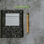 A black and white composition notebook, two yellow pencils and a green highlighter laid out on a gray concrete surface