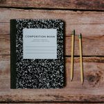 A black-and-white composition notebook and two sharpened pencils on a weathered wooden table, seen from above