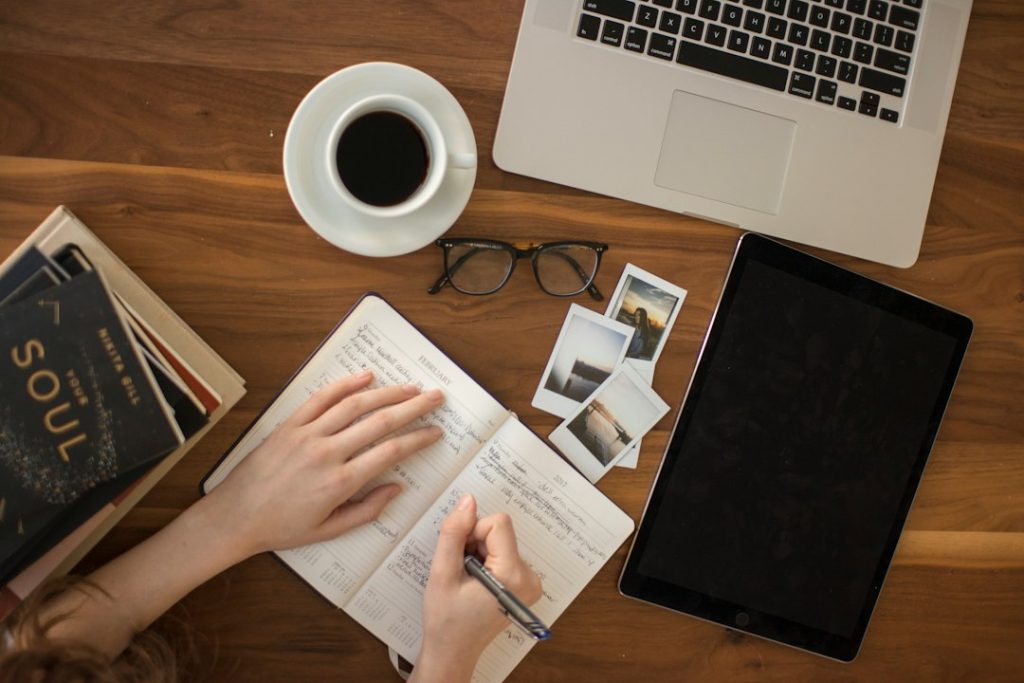 Overhead view of a desk: a person writing in a paper planner beside a coffee cup, glasses, instant photos, a laptop and a tablet