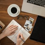 Overhead view of a desk: a person writing in a paper planner beside a coffee cup, glasses, instant photos, a laptop and a tablet