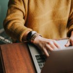 A person in a mustard knit sweater and gold bracelets typing on a laptop at a wooden desk
