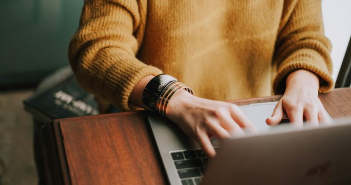 A person in a mustard knit sweater and gold bracelets typing on a laptop at a wooden desk