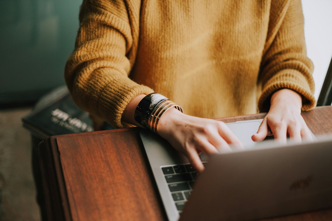 A person in a mustard knit sweater and gold bracelets typing on a laptop at a wooden desk