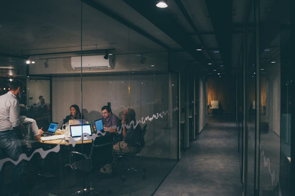 Four people working together around a small table with laptops in a glass-walled office at night