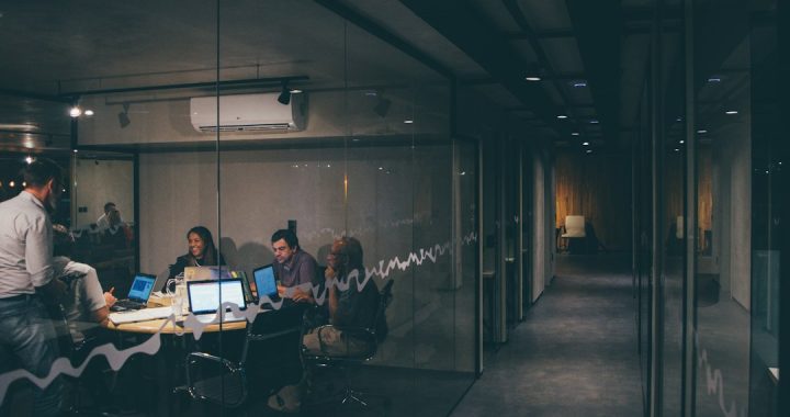 Four people working together around a small table with laptops in a glass-walled office at night