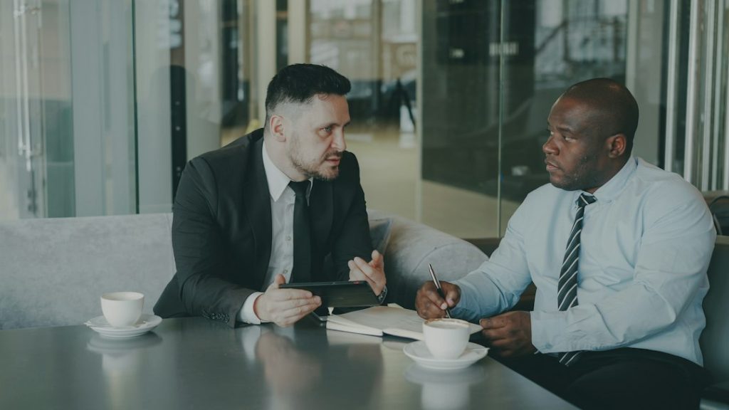 Two men in business attire talking across a cafe table, one holding a tablet, the other taking notes with a pen