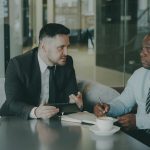 Two men in business attire talking across a cafe table, one holding a tablet, the other taking notes with a pen