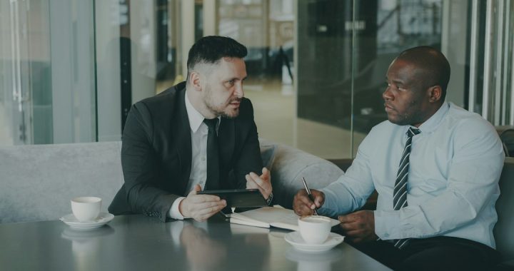 Two men in business attire talking across a cafe table, one holding a tablet, the other taking notes with a pen