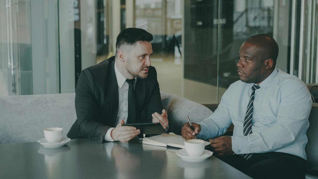Two men in business attire talking across a cafe table, one holding a tablet, the other taking notes with a pen