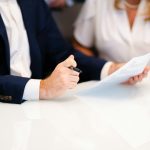 Two people at a white table reviewing a printed document, one holding a pen over the page