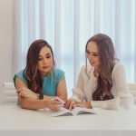 Two women with long hair sitting at a white desk in a bright room, looking together at an open book while one holds a pink highlighter