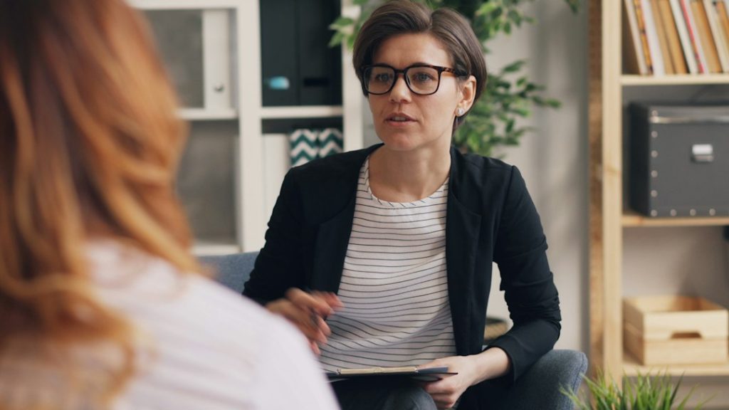 A woman with short hair and glasses holding a clipboard, listening during a one-on-one session with a client whose back is to the camera