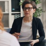 A woman with short hair and glasses holding a clipboard, listening during a one-on-one session with a client whose back is to the camera