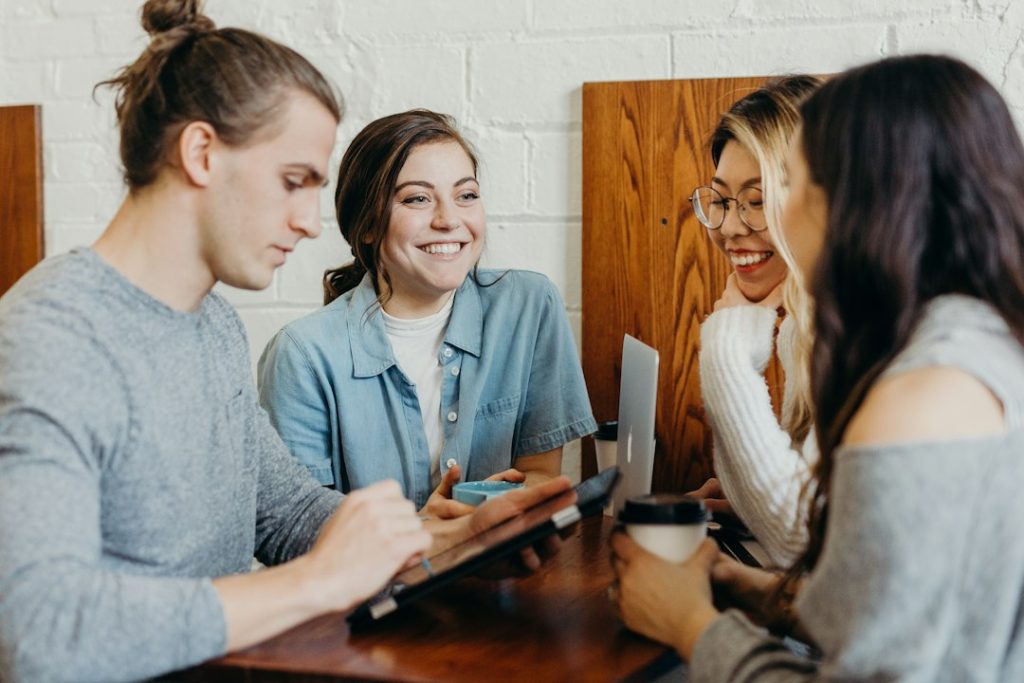 Four people sitting around a wooden table against a white brick wall, smiling and talking while one of them holds a tablet, with coffee cups and an open laptop nearby