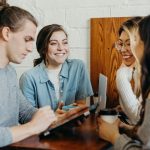 Four people sitting around a wooden table against a white brick wall, smiling and talking while one of them holds a tablet, with coffee cups and an open laptop nearby