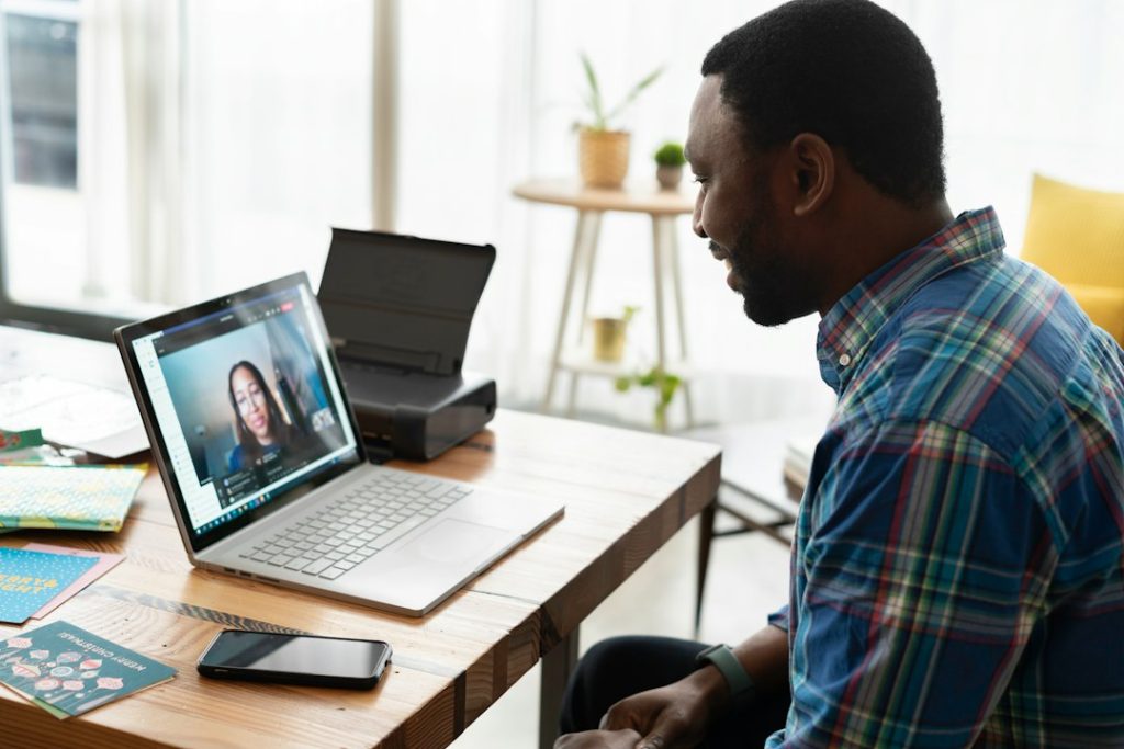 A man in a plaid shirt smiles at a laptop on a wooden desk, on a video call with a woman, with a phone and greeting cards beside him.