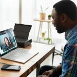 A man in a plaid shirt smiles at a laptop on a wooden desk, on a video call with a woman, with a phone and greeting cards beside him.