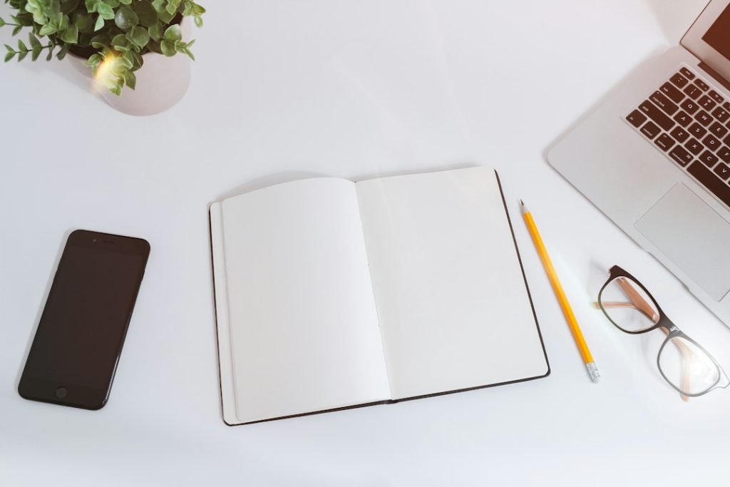 An open blank notebook with a yellow pencil, reading glasses, a phone and a laptop corner on a white desk beside a green plant