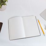 An open blank notebook with a yellow pencil, reading glasses, a phone and a laptop corner on a white desk beside a green plant
