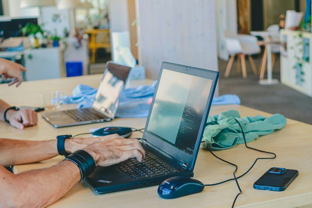 A person typing on a laptop at a light wooden desk in a bright shared workspace, with a second laptop and a phone beside it.