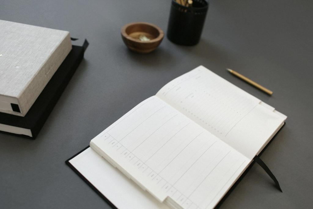 An open weekly planner with blank ruled columns on a dark grey desk, next to a gold pencil, a stack of linen-covered notebooks, a small wooden bowl and a black pen cup.