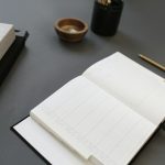An open weekly planner with blank ruled columns on a dark grey desk, next to a gold pencil, a stack of linen-covered notebooks, a small wooden bowl and a black pen cup.